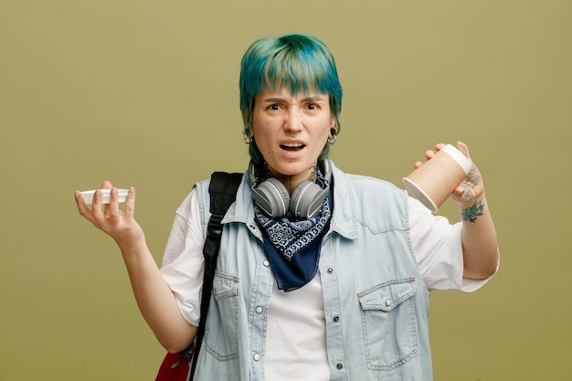 Annoyed young female student wearing headphones and bandana on neck and backpack holding paper coffee cup upside down and its cap looking at camera isolated on olive green background
