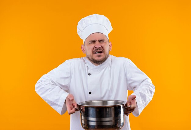 Annoyed young handsome cook in chef uniform holding boiler with closed eyes on isolated orange wall