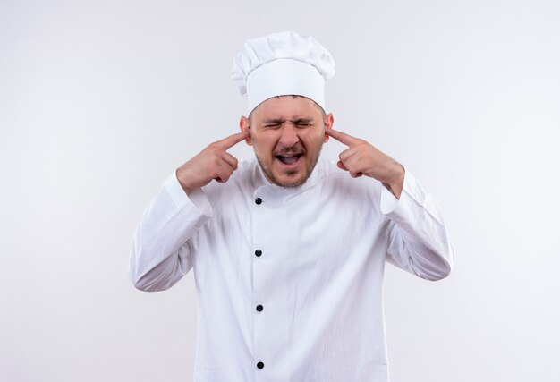 Annoyed young handsome cook in chef uniform putting fingers in ears with closed eyes isolated on white wall