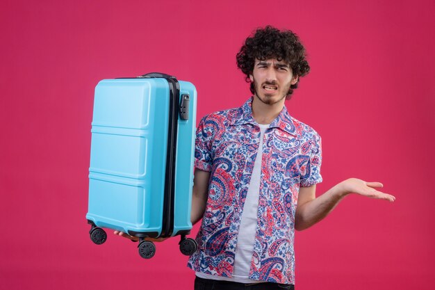 Annoyed young handsome curly traveler man holding suitcase and showing empty hand on isolated pink space