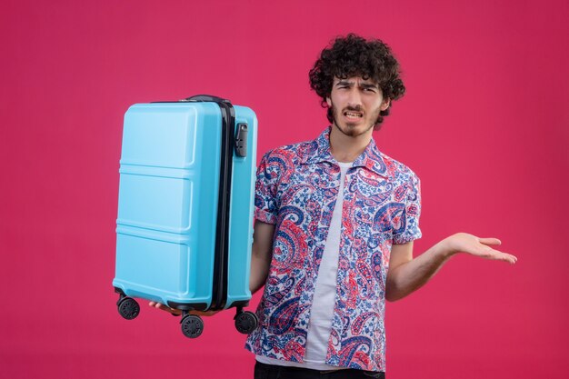 スーツケースを持って孤立したピンクのスペースに空の手を見せてイライラする若いハンサムな巻き毛の旅行者の男