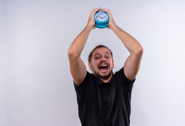 Annoyed young handsome man in black polo shirt holding alarm clock going to throw and break it standing over white background