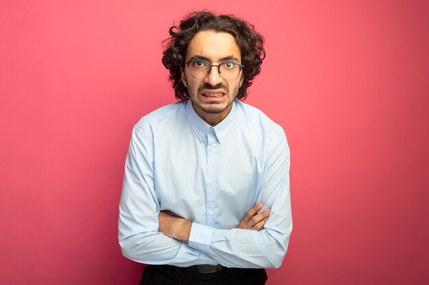 Annoyed young handsome man wearing glasses standing with closed posture looking at front isolated on pink wall