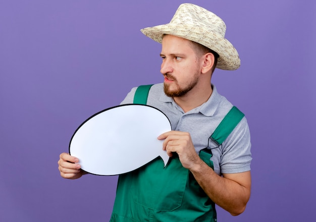 Infastidito giovane giardiniere slavo bello in uniforme e cappello tenendo la bolla di chat guardando il lato isolato sulla parete viola