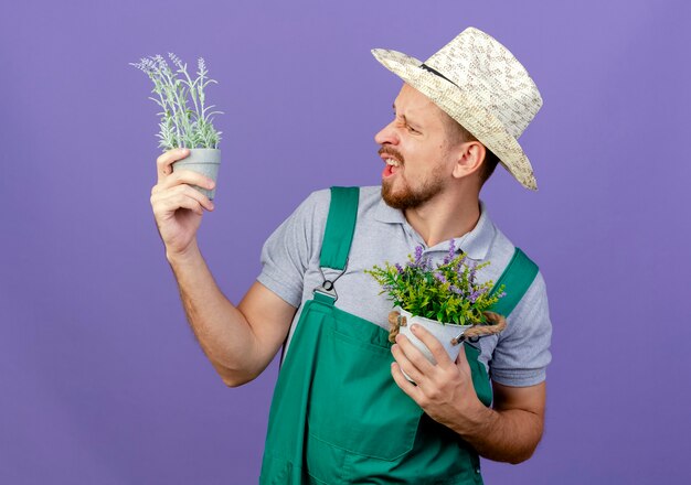 コピースペースで紫色の壁に隔離された植木鉢を保持し、見て帽子と制服を着たイライラした若いハンサムなスラブの庭師