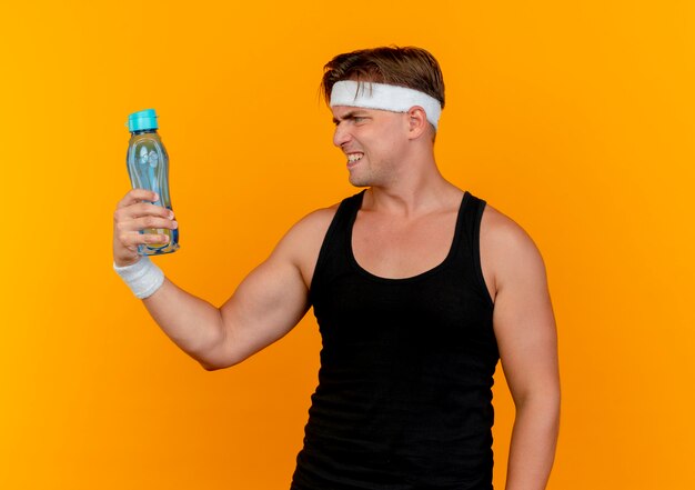 Annoyed young handsome sporty man wearing headband and wristbands holding and looking at water bottle isolated on orange 