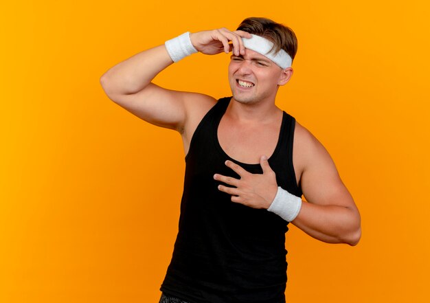 Annoyed young handsome sporty man wearing headband and wristbands looking at side putting hands on forehead and on chest isolated on orange 