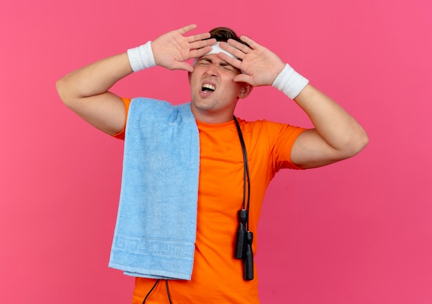 Free photo annoyed young handsome sporty man wearing headband and wristbands with towel and jump rope around neck keeping hands near face with closed eyes isolated on pink