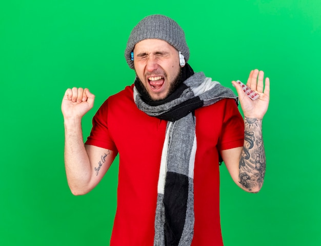 Annoyed young ill man wearing and holding packs of medical pills under winter hat and wearing scarf keeps fist and holds pack of medical pills isolated on green wall