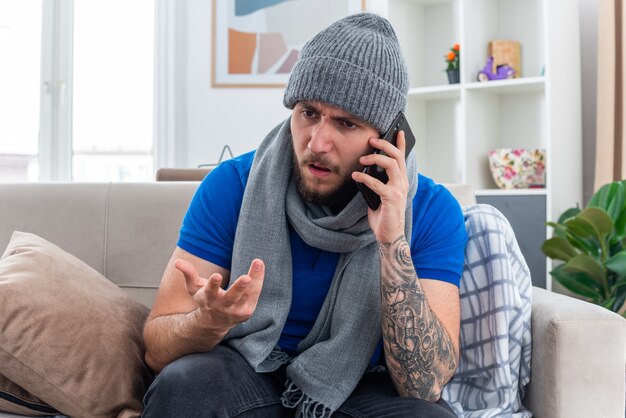раздраженный молодой больной человек в шарфе и зимней шапке сидит на диване в гостиной, глядя в сторону, разговаривает по телефону, показывая пустую руку