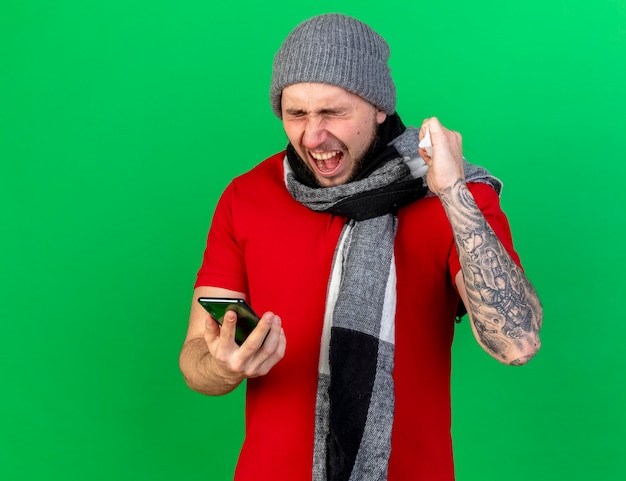 Annoyed young ill man wearing winter hat and scarf keeps fist holding tissue and holds phone isolated on green wall