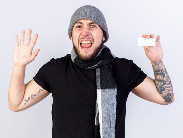 Annoyed young ill man wearing winter hat and scarf stands with raised hand holding pack of medical tablets isolated on white wall