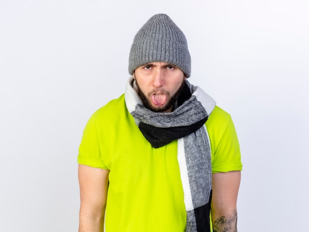 Annoyed young ill man wearing winter hat and scarf stucks out tongue isolated on white wall