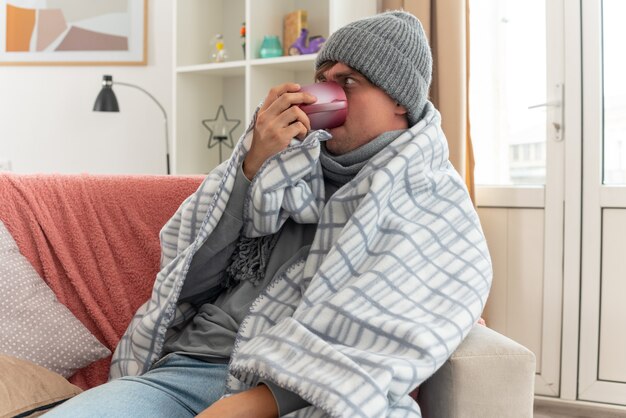 annoyed young ill man with scarf around neck wearing winter hat wrapped in plaid drinking from cup sitting on couch at living room