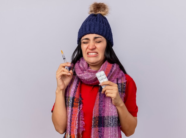 Free photo annoyed young ill woman wearing winter hat and scarf holding syringe and pack of tablets with closed eyes isolated on white wall