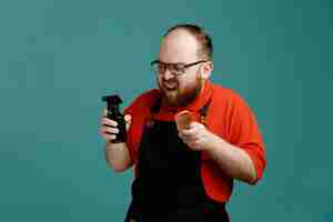 Free photo annoyed young male barber wearing glasses red shirt and barber apron holding hair spray and comb looking at side isolated on blue background