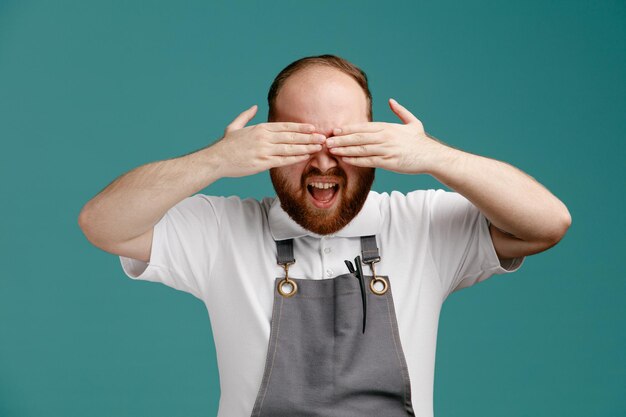 Annoyed young male barber wearing white shirt and barber apron keeping hands in front of eyes isolated on blue background