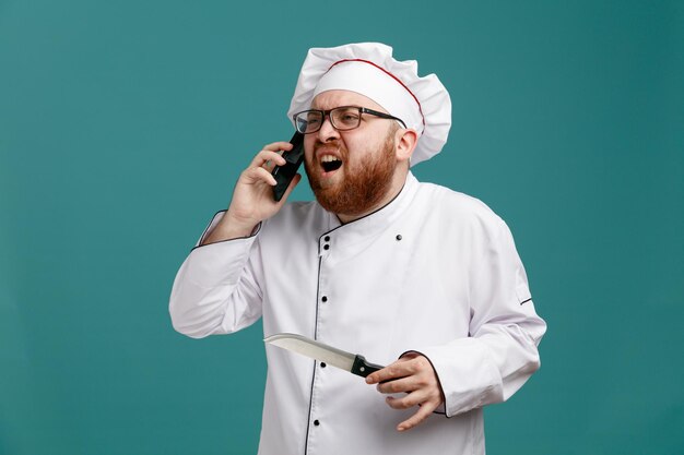 Annoyed young male chef wearing glasses uniform and cap holding knife looking at side talking on phone isolated on blue background