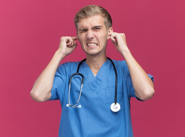 ピンクの壁に分離された聴診器の耳を閉じる医者の制服を着てイライラする若い男性医師
