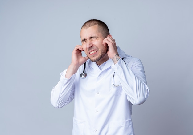 Annoyed young male doctor wearing medical robe and stethoscope around his neck putting fingers in ears with closed eyes isolated on white  with copy space