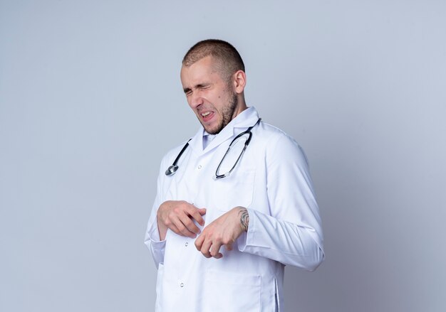 Annoyed young male doctor wearing medical robe and stethoscope around his neck putting hand on belly with closed eyes isolated on white  with copy space