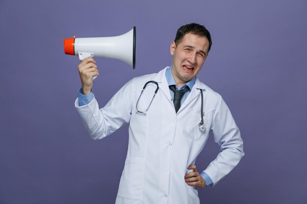 Annoyed young male doctor wearing medical robe and stethoscope around neck keeping hand on waist looking at camera holding speaker pointing it at himself isolated on purple background