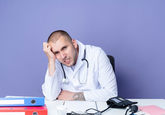 Раздраженный молодой мужчина-врач в медицинском халате и стетоскопе сидит за столом с рабочими инструментами, положив руку на голову, изолированную на фиолетовом