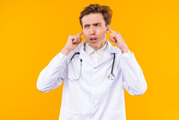 Annoyed young male doctor wearing medical robe with stethoscope closed ears isolated on orange wall
