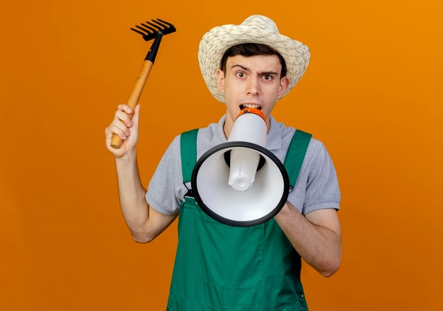 Annoyed young male gardener wearing gardening hat holds rake speaks into loud speaker 