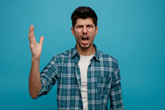Annoyed young man looking at camera keeping hand in air isolated on blue background
