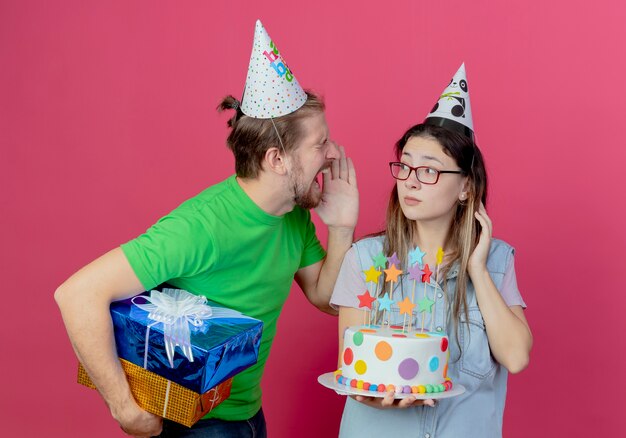 Annoyed young man wearing party hat holds gift boxes looking and shouting at young girl wearing party hat and holding birthday cake isolated on pink wall