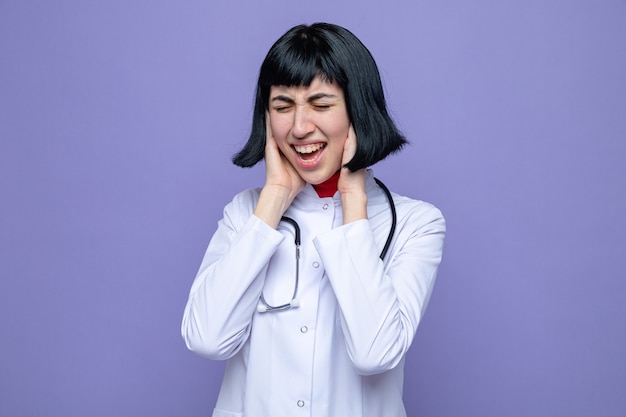 Infastidita giovane bella ragazza caucasica in uniforme da medico con uno stetoscopio che si copre le orecchie con le mani