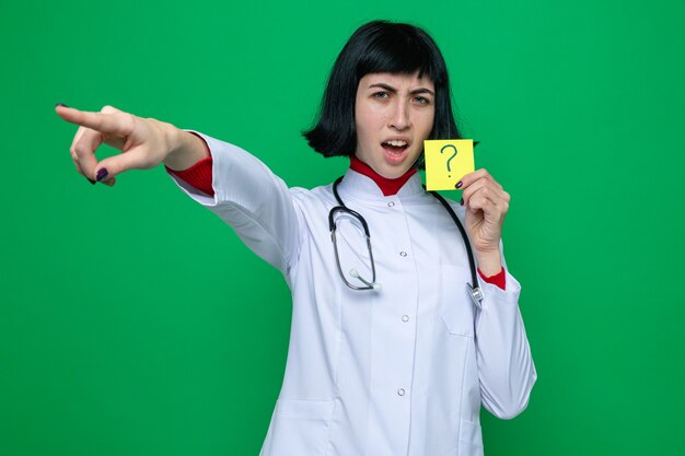 Раздраженная молодая симпатичная кавказская девушка в униформе врача со стетоскопом держит желтую карточку со знаком вопроса и указывает в сторону