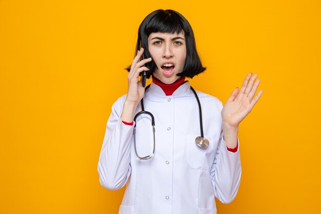 Раздраженная молодая симпатичная кавказская девушка в медицинской форме со стетоскопом разговаривает по телефону и держит руку открытой