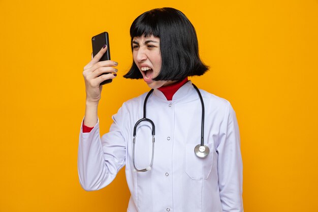 Раздраженная молодая симпатичная кавказская девушка в медицинской форме со стетоскопом кричит на кого-то по телефону