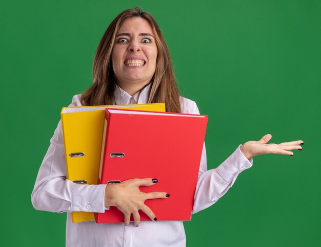 Annoyed young pretty caucasian girl holds file folders and points at side with hand isolated on green wall with copy space