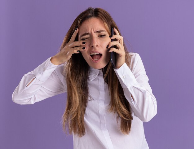 イライラする若いかなり白人の女の子が電話で話し、コピースペースで紫色の壁に隔離された顔に手を置きます