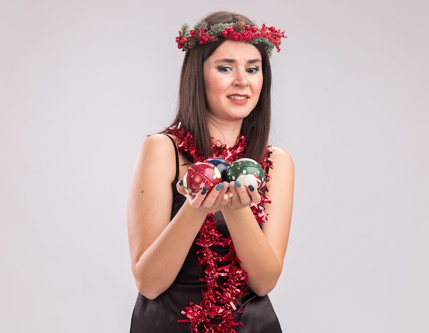 Annoyed young pretty caucasian girl wearing christmas head wreath and tinsel garland around neck holding and looking at christmas baubles isolated on white background with copy space