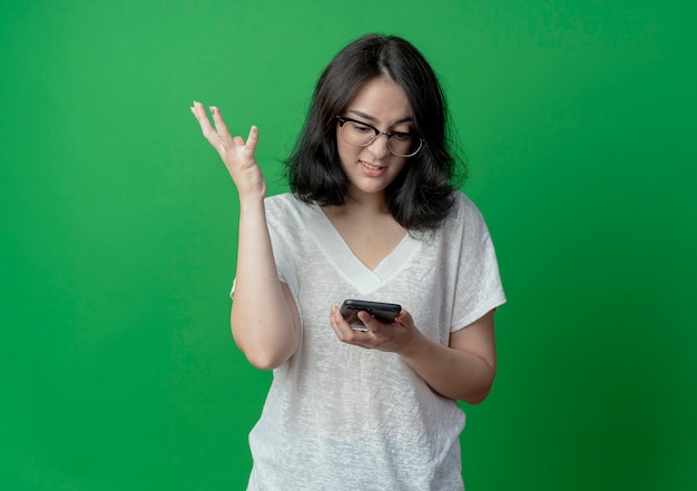Annoyed young pretty caucasian girl wearing glasses holding and looking at mobile phone and keeping hand in air isolated on green background with copy space