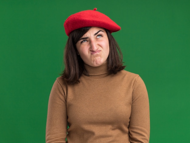 Foto gratuita ragazza abbastanza caucasica giovane infastidita con il cappello del berretto che cerca sul verde