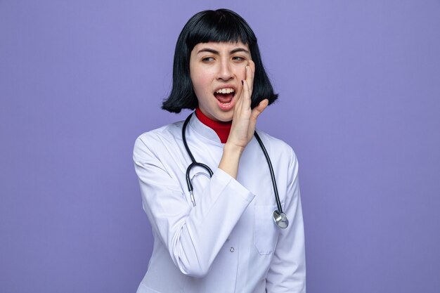 Annoyed young pretty caucasian woman in doctor uniform with stethoscope keeping hand close to her mouth and looking at side 