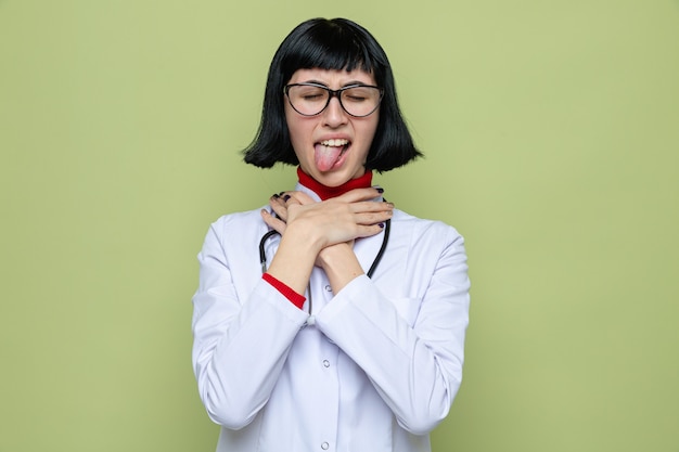 Раздраженная молодая симпатичная кавказская женщина в очках в медицинской форме со стетоскопом, высунув язык и задыхаясь руками