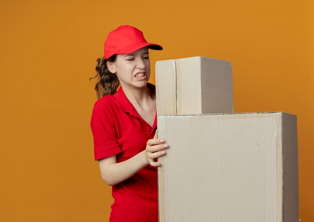 赤い制服と帽子を保持し、コピースペースでオレンジ色の背景に分離されたカートンボックスを見てイライラする若いかわいい配達の女の子