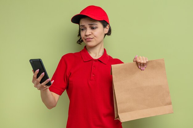 Annoyed young pretty delivery woman holding paper food packaging and looking at phone 