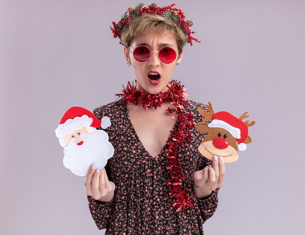 Free photo annoyed young pretty girl wearing christmas head wreath and tinsel garland around neck with glasses holding christmas reindeer and santa claus paper ornaments  isolated on white wall