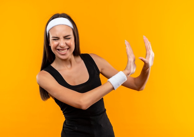 Раздраженная молодая симпатичная спортивная девушка с повязкой на голову и браслетом, жестом показывающая нет, с закрытыми глазами и вытянутыми руками, изолированными на оранжевой стене