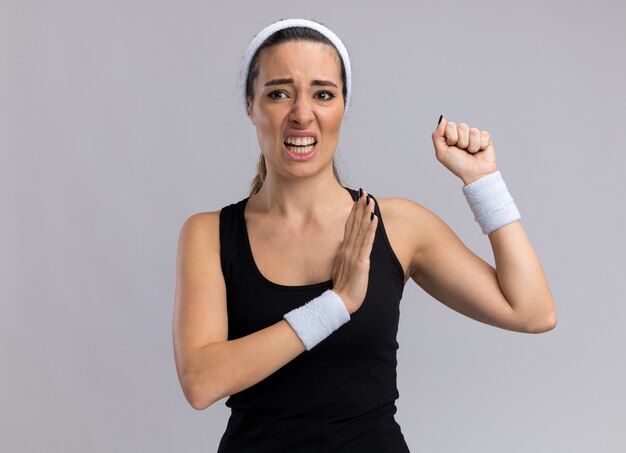 Annoyed young pretty sporty girl wearing headband and wristbands looking at side clenching fist keeping hand in air 