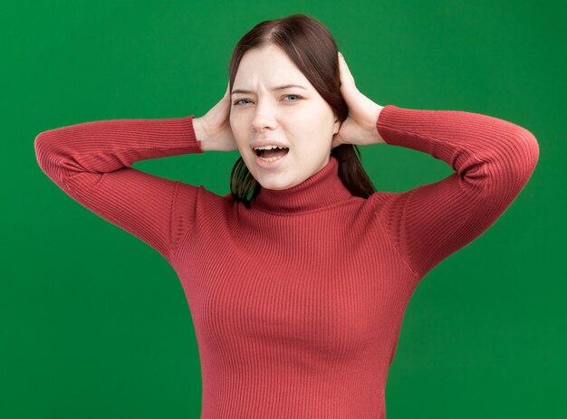 Annoyed young pretty woman looking at front putting hands on head isolated on green wall