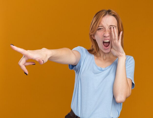 Free photo annoyed young redhead ginger girl with freckles holding hand close to mouth and pointing at side on orange