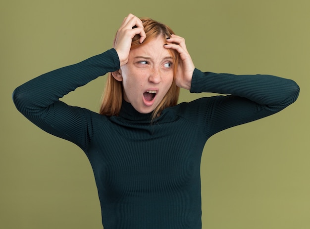 Annoyed young redhead ginger girl with freckles puts hands on head and looks at side isolated on olive green wall with copy space